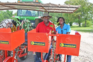 Warren Dixon sits to the left of Sabrenna Bryant on seats on the back of a transplanting machine that is hooked up to a tractor.