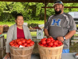 Rosalina Nuñez stands on the right and Andy Ordaz on the left behind two bushel baskets of tomatoes on a table.