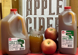 A mason jar full of apple cider and two apples sit in between two jugs of apple cider. An apple cider sign in the background.