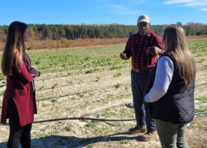 Two women stand in the foreground with a gentleman in the background, standing in a field.