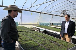 Inside a greenhouse, Zach Ducheneaux stands to the left of a table with growing microgreens and Eric McClam stands on the right of the table.