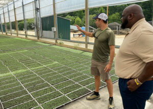 Ron Howell stands in the foreground and Eric McClam shows him growing microgreens inside a greenhouse.